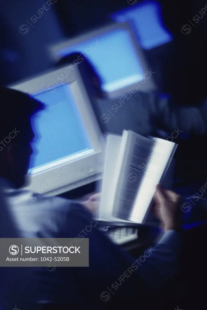 Rear view of a man reading a book in front of a computer monitor