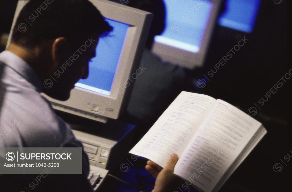 Stock Photo: 1042-5208 Rear view of a young man reading a book in front of a computer monitor