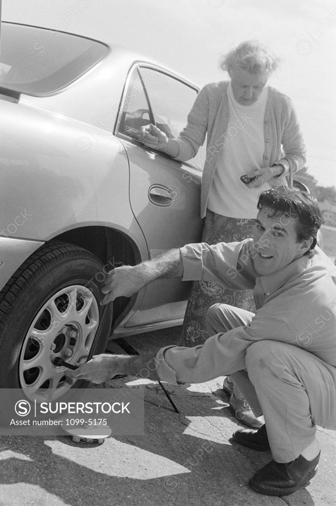 Stock Photo: 1099-5175 Senior woman standing beside her car with a mid adult man changing a flat tire