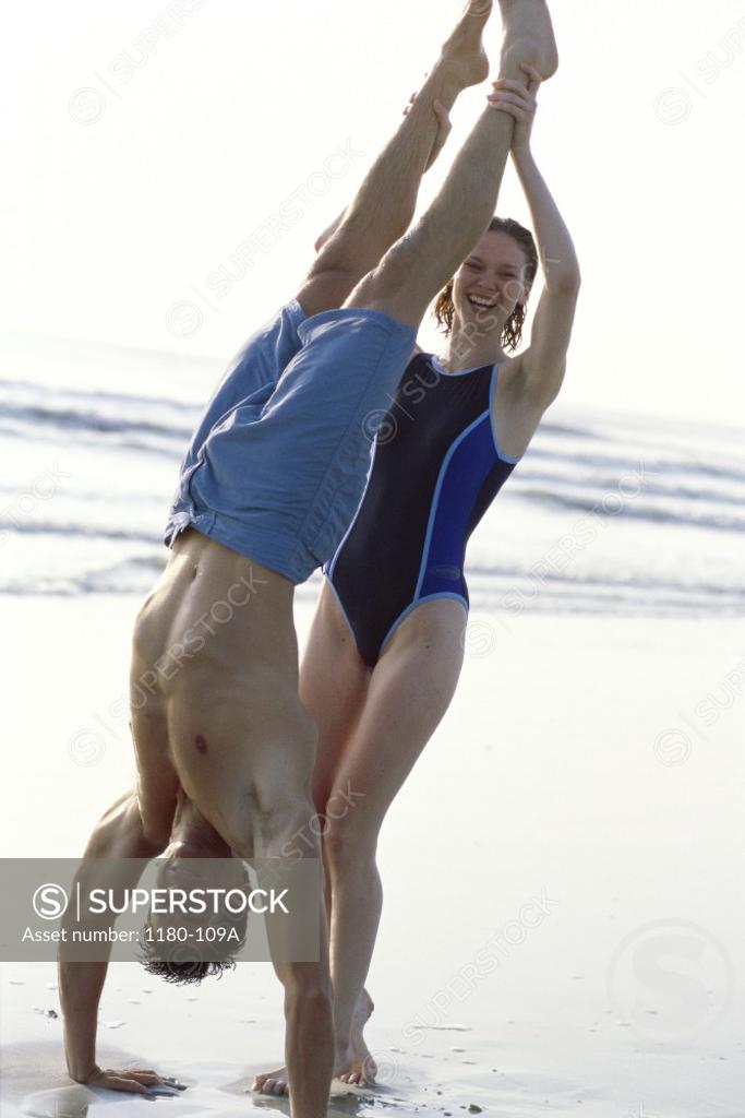 Stock Photo: 1180-109A Young man doing a hand stand on the beach with a young woman supporting him