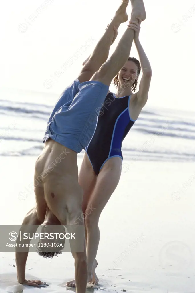 Young man doing a hand stand on the beach with a young woman supporting him