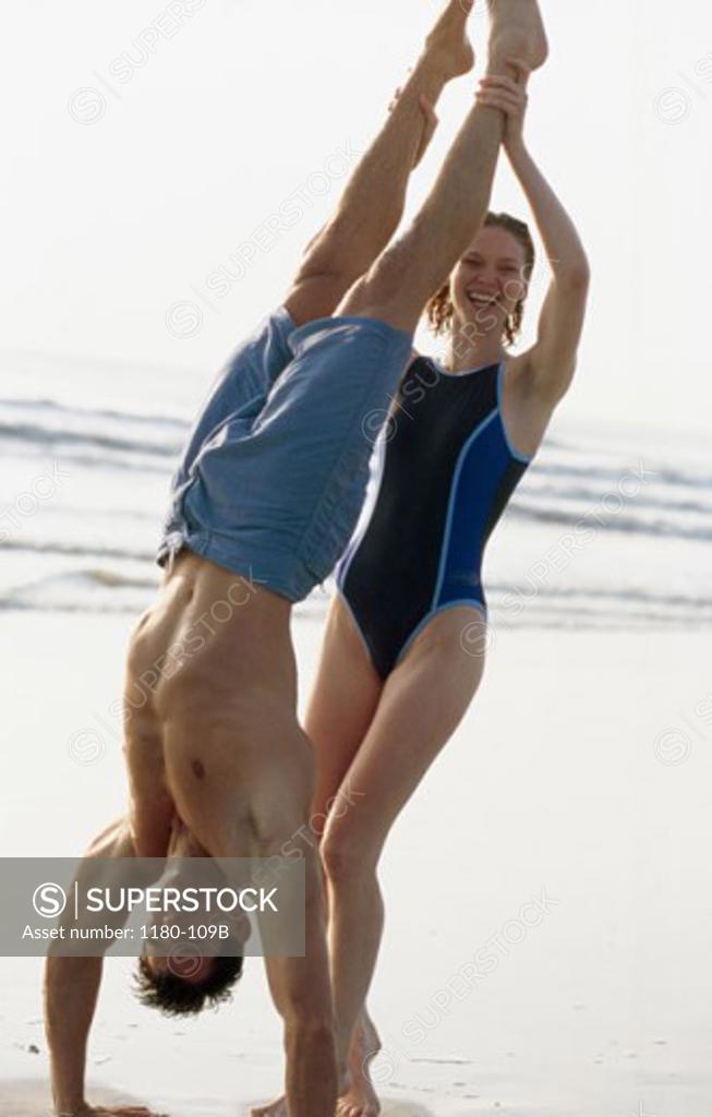 Stock Photo: 1180-109B Young man doing a hand stand on the beach with a young woman supporting him