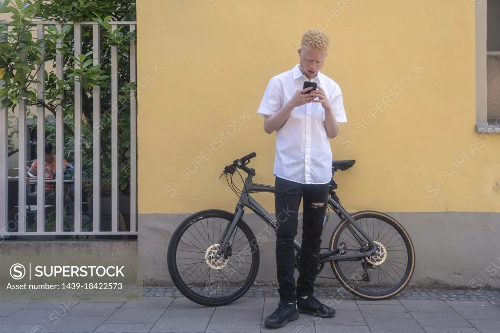 Germany, Cologne, Albino man using smart phone in front of building and bicycle