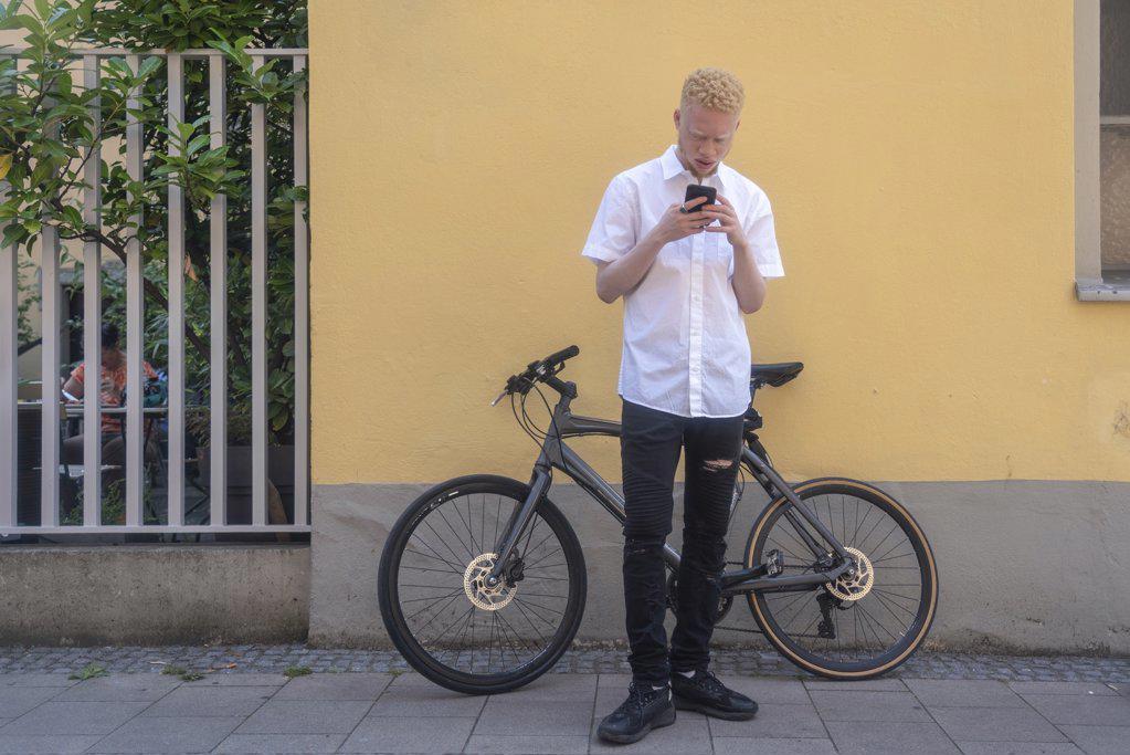 Germany, Cologne, Albino man using smart phone in front of building and bicycle