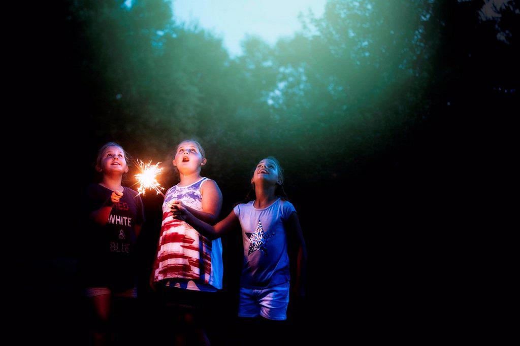 Three girls holding sparklers looking up from garden at night on independence day, USA