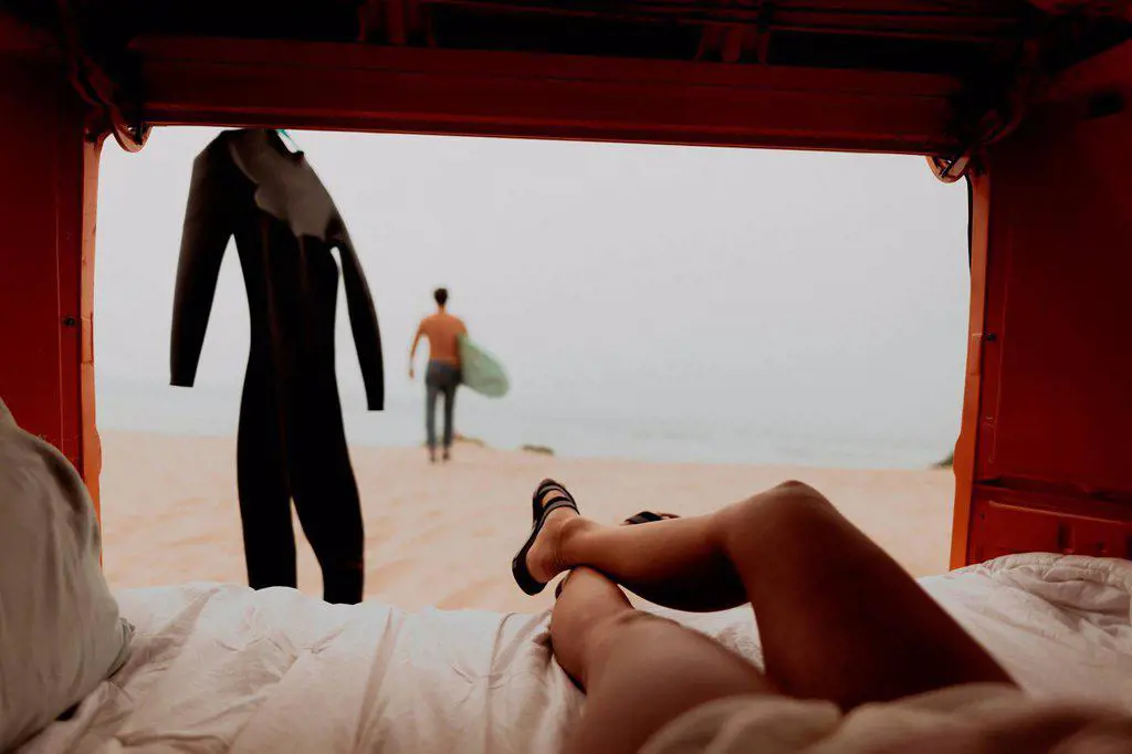 Young woman reclining in back of recreational vehicle watching surfer boyfriend on beach, Jalama, Ventura, California, USA