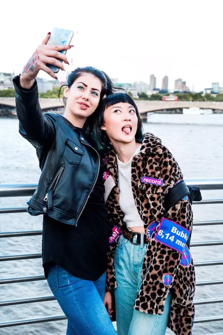 Two young stylish women taking smartphone selfie on millennium footbridge, London, UK