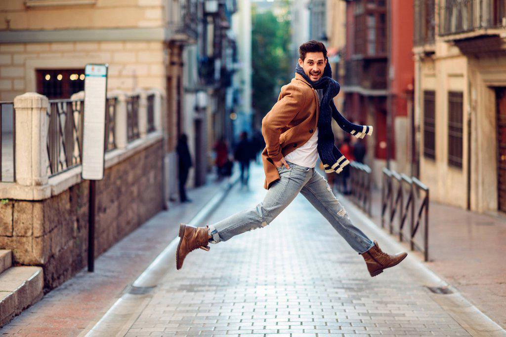 Young happy man jumping wearing winter clothes in the street. Young bearded guy with modern hairstyle with coat, scarf, blue jeans and t-shirt in urba...