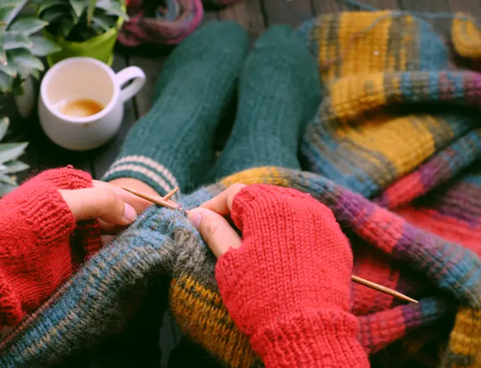 Top view woman feet with socks, sitting at home balcony, hand with knitted gloves hold knitting needle to knit colorful wool scarf  for meaningful handmade winter gift when wintertime come.