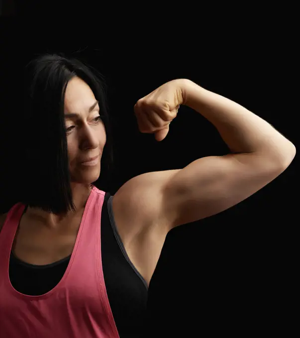 beautiful athletic girl raised and bent her arm demonstrating her biceps, athlete is standing on a dark background, wearing a black top 