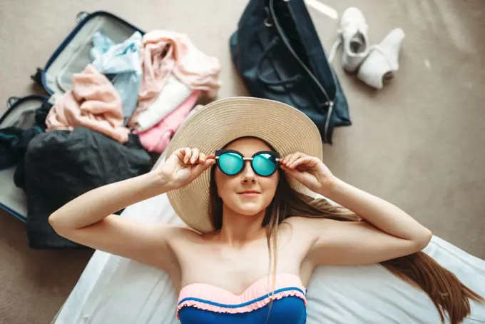 Young woman lies in swimsuit and sunglasses, top view, opened suitcase with clothes on background. Fees on journey concept. Preparation for summer vacation. Woman lies in swimsuit and sunglasses, top view