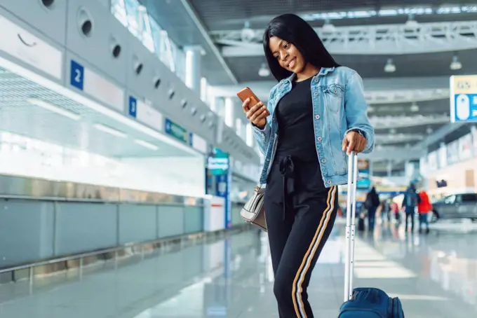 Black female tourist with suitcase and phone waiting for departure in international airport. Passenger with baggage in air terminal, happy journey sexy lady, summer travel of happy woman