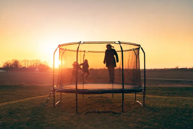 Trampoline on a lawn in the sunset with two kids and a young woman juping and playing