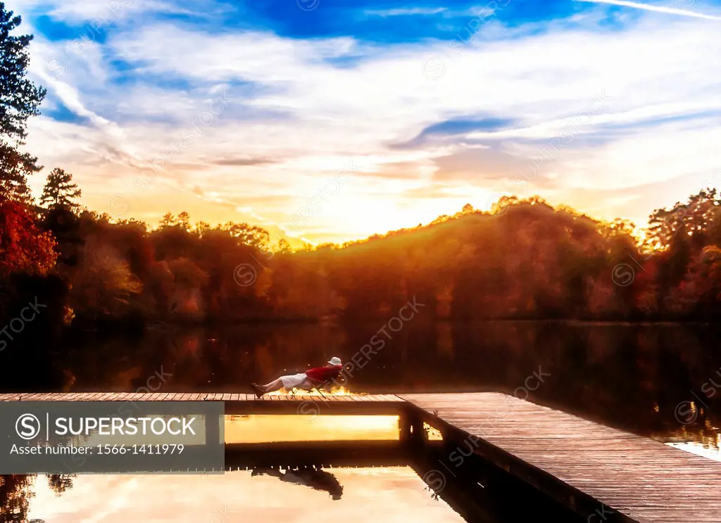 A 53 year old man lounging on a folding chair on a wooden dock by a lake with fall foliage in the background. Oak Mountain State Park, Alabama.