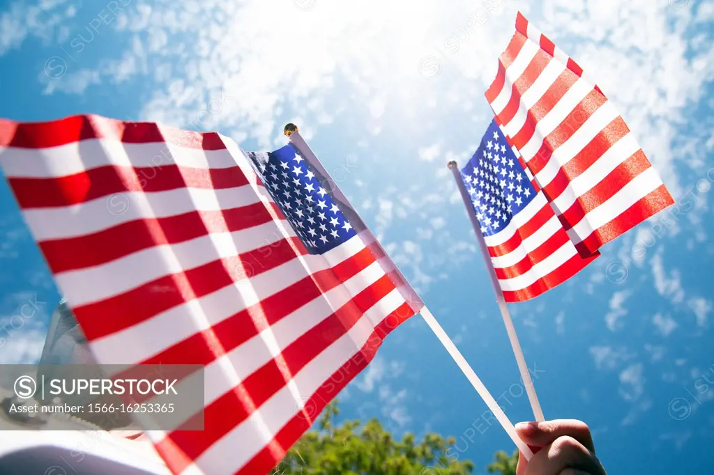 Hand holding two american flags on the blue sky with sunlight background, waving flag for United States of America close-up beauty.