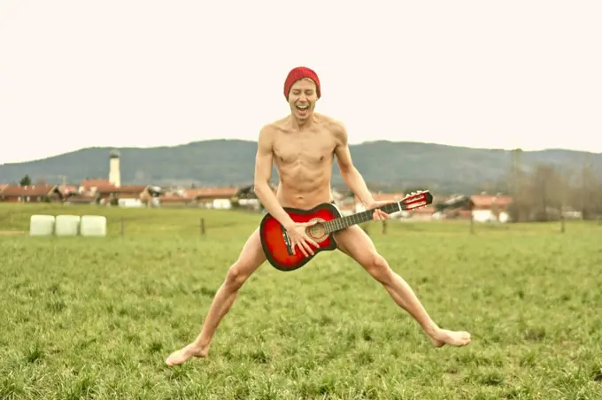 naked young man jumping on countryside meadow with guitar, musician, rebel, naked, in Waakirchen, Bavaria, Germany