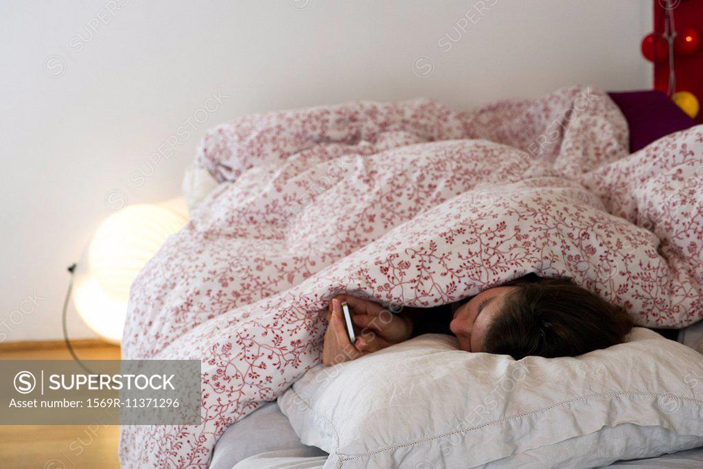Stock Photo: 1569R-11371296 Woman lying in bed under covers, looking at smartphone