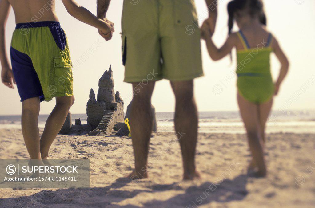 Stock Photo: 1574R-014541E Rear view of a father holding his children's hands looking at a sand castle on the beach