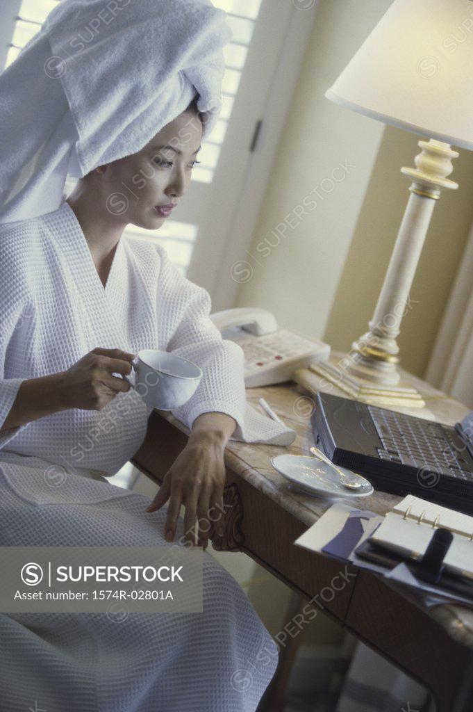 Stock Photo: 1574R-02801A Young woman wearing a bathrobe holding a cup of coffee and looking at a laptop in a hotel room