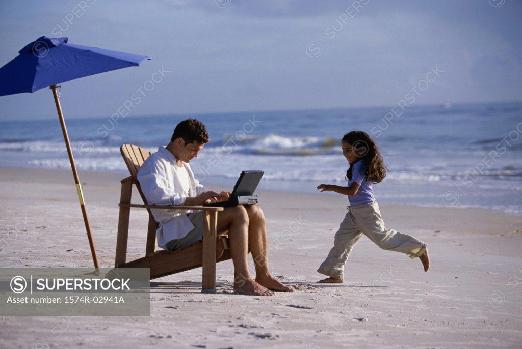 Stock Photo: 1574R-02941A Father working on a laptop with his daughter running on the beach