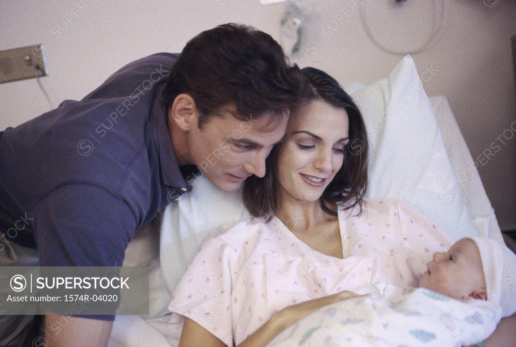 Stock Photo: 1574R-04020 Mother lying in bed holding her baby boy with father standing beside them