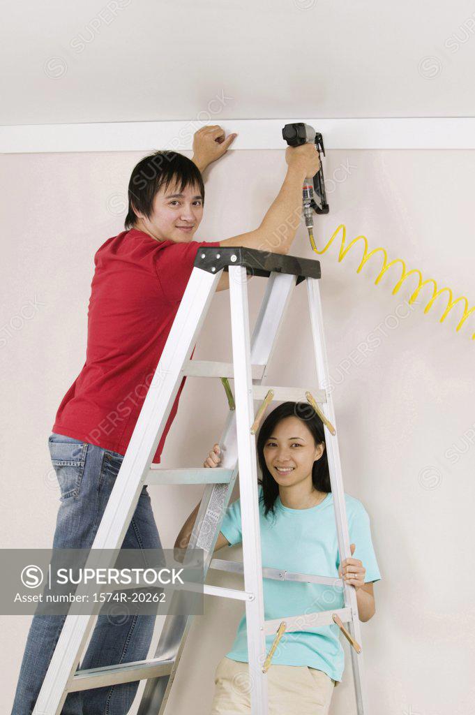 Stock Photo: 1574R-20262 Portrait of a young man using a drill on a wall with a young woman holding a ladder