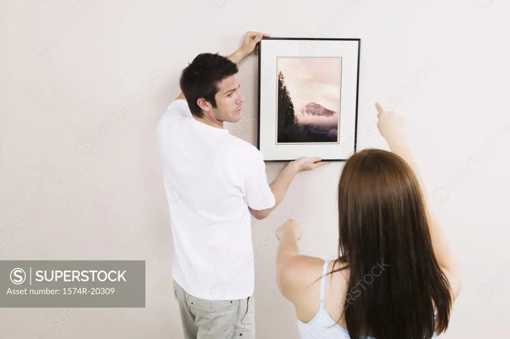 Rear view of a young man putting up a painting on a wall with a young woman's guidance