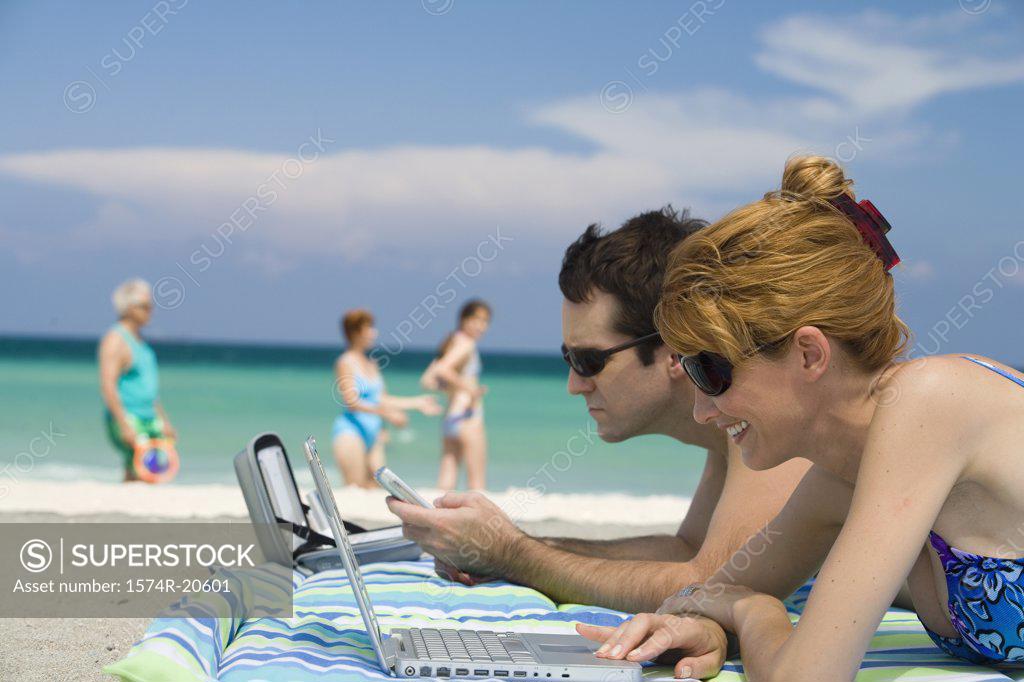 Stock Photo: 1574R-20601 Side profile of a young woman using a laptop with a young man lying beside her looking at a mobile phone