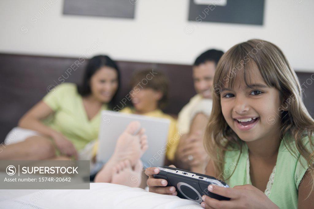 Stock Photo: 1574R-20642 Portrait of a girl holding a handheld video game with her parents and brother looking at a laptop in the background
