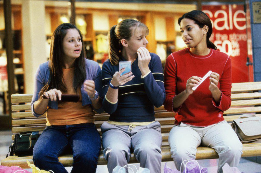 Three teenage girls sitting on a bench in a shopping mall applying make-up