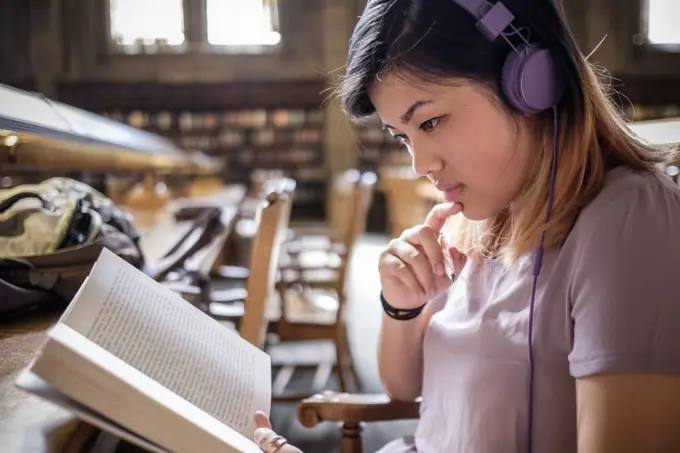 Chinese woman in library reading book and listening to headphones
