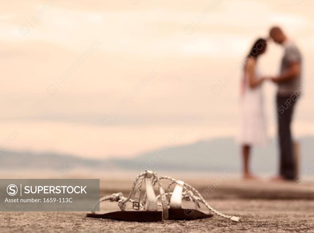 Stock Photo: 1659-113C Close-up of a pair of sandals with a young couple standing in the background on the beach
