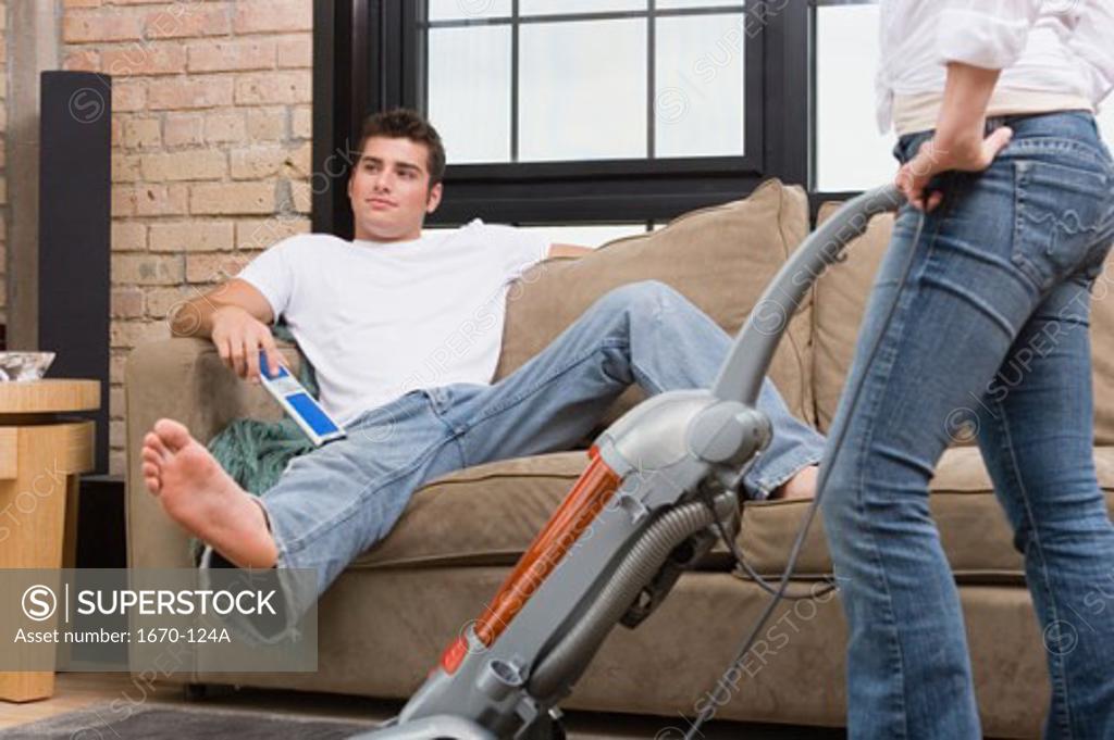Stock Photo: 1670-124A Teenage boy sitting on a couch with a woman vacuuming the floor