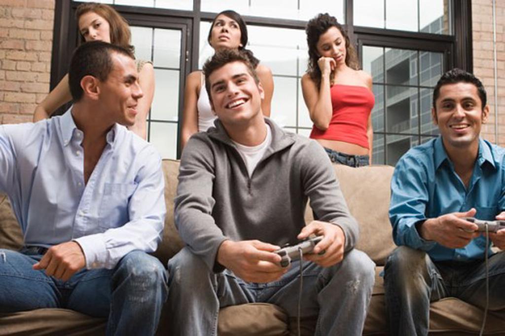 Young man and a teenage boy playing a video game with three young women standing behind them looking displeased