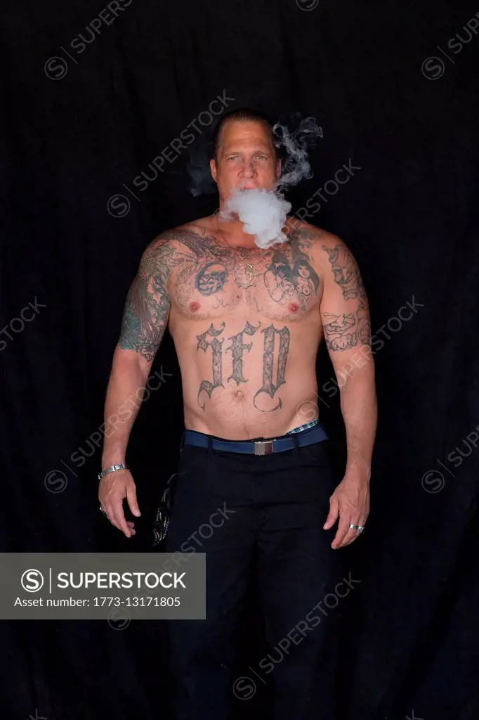 Front view of bare chested tattooed mature man, hair slicked back looking at camera blowing smoke