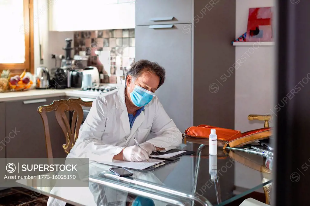 Doctor wearing a white coat, facemask and protective gloves sitting at a kitchen table writing up medical notes.