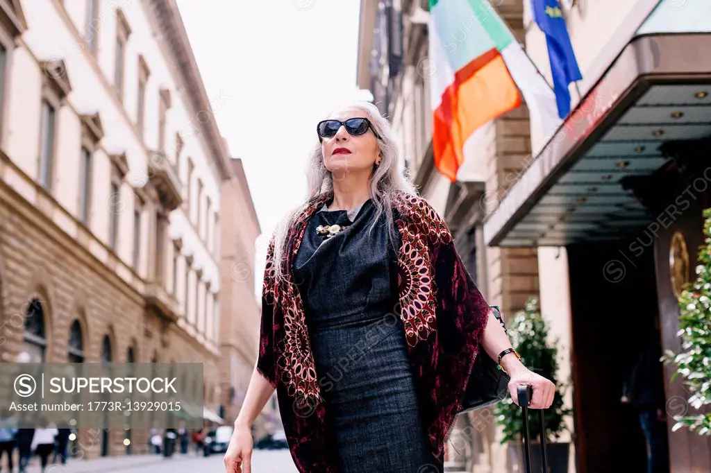 Mature woman with long grey hair with wheeled suitcase outside hotel in Florence, Italy