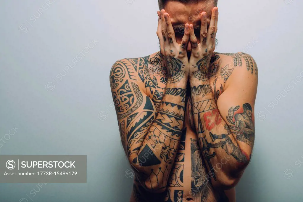 Portrait of young man with beard, covered in tattoos, hands covering face -  SuperStock