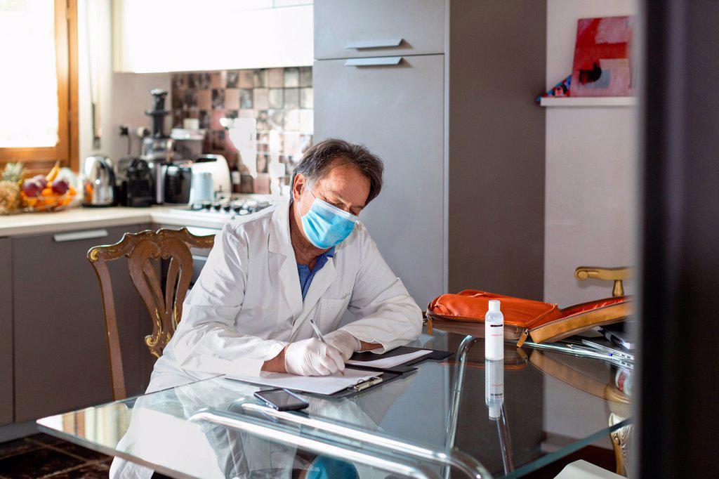 Doctor wearing a white coat, facemask and protective gloves sitting at a kitchen table writing up medical notes.