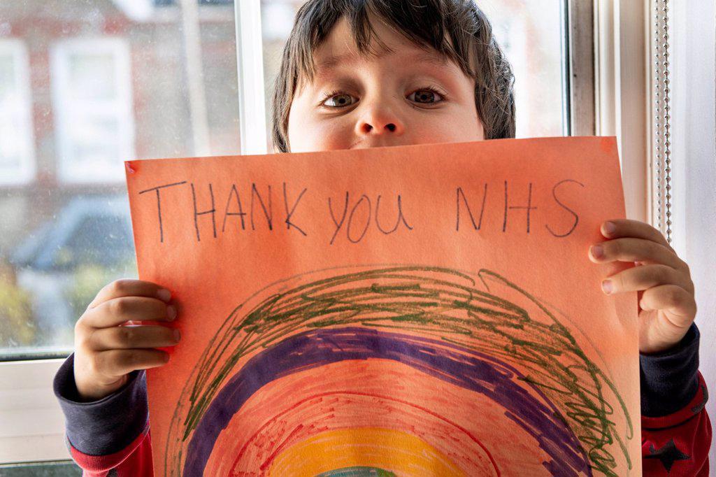 A boy holding up a drawing of a rainbow with the words Thank You NHS