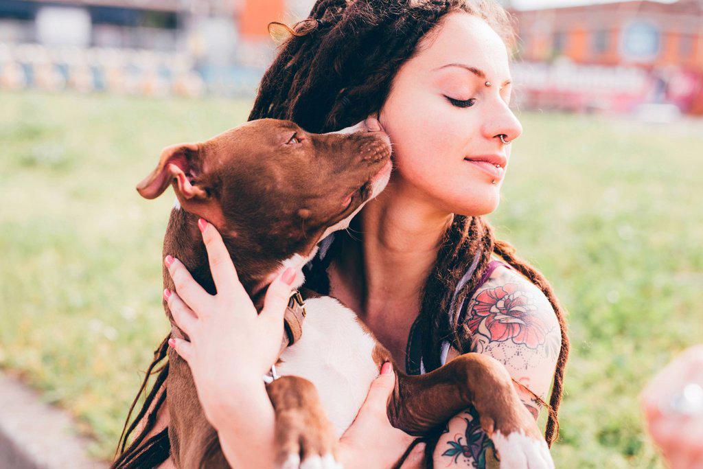 Pit bull terrier licking tattooed young woman in urban park