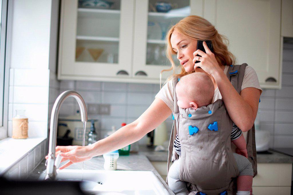 Woman holding baby boy in sling, using smartphone, turning on tap to do washing up