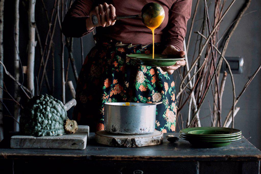 Young woman ladling fresh soup from saucepan at rustic kitchen counter, mid section