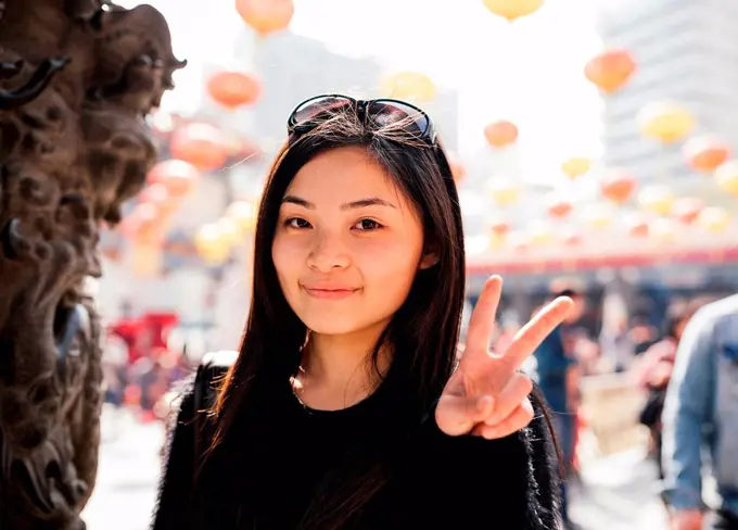 Portrait of young woman with long hair and sunglasses on head doing peace sign, looking at camera