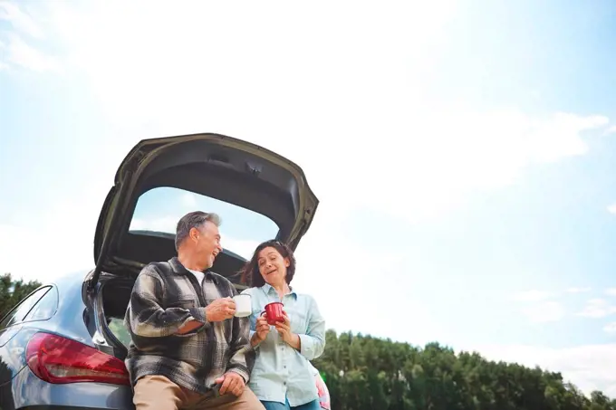 Mature couple standing beside car, holding tin cups, smiling, low angle view