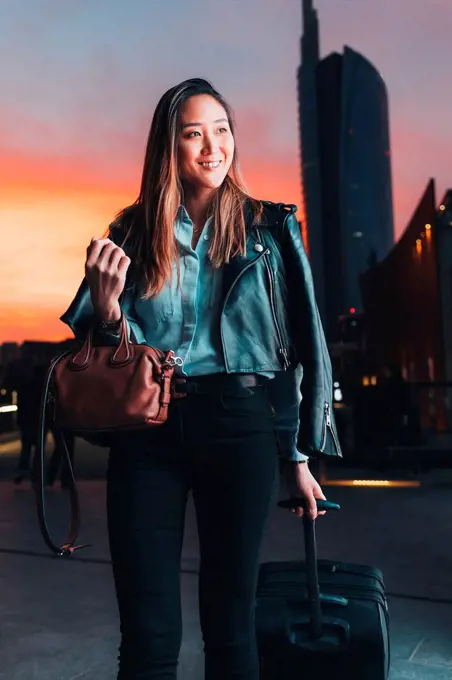 Businesswoman walking outdoors, at sunset, pulling wheeled suitcase, smiling