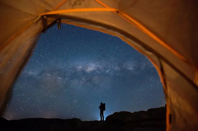Silhouette of backpacker standing against starry night sky and Milky Way galaxy visible from inside of pitched tent, New South Wales, Australia
