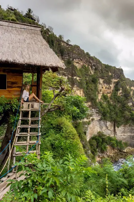 Woman sitting outside thatched roof hut on coast with cliffs, Nusa Penida, Bali, Indonesia