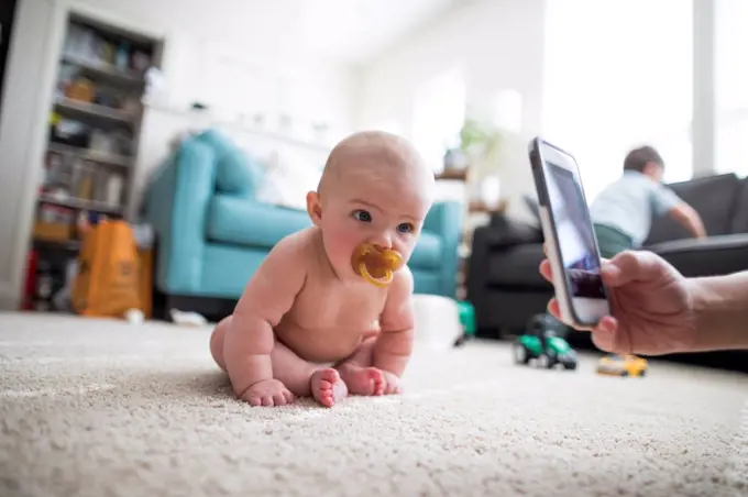Parent photographing baby sitting on floor with smartphone