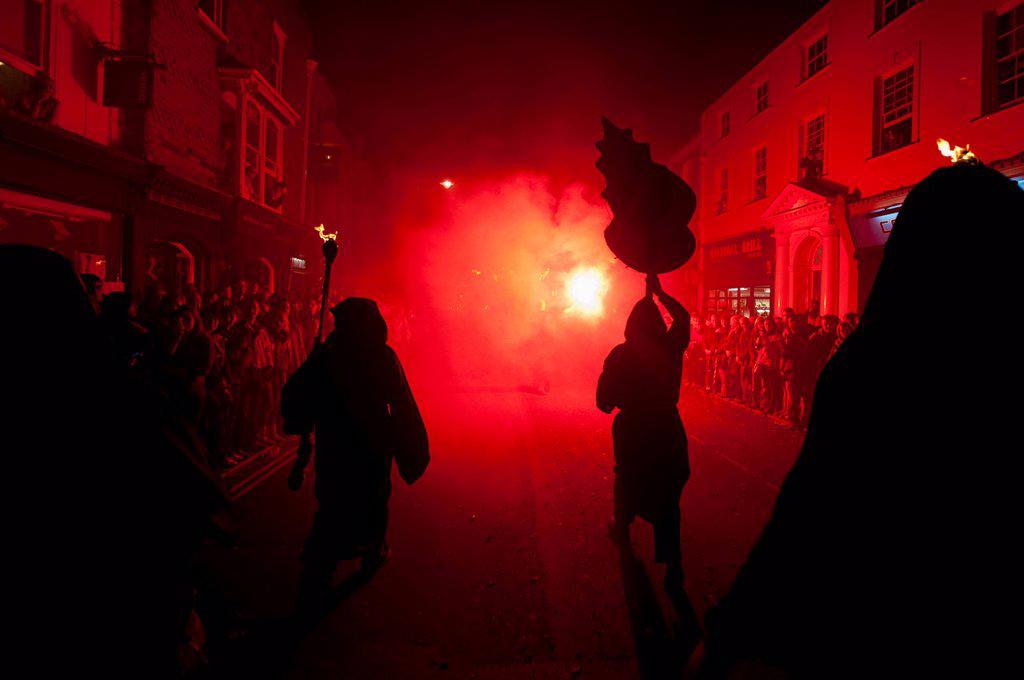 Silhouettes of monks from Southover Bonfire Society lit by red flares on United Grand Procession down High Street, Bonfire Night; Lewes, East Sussex, ...